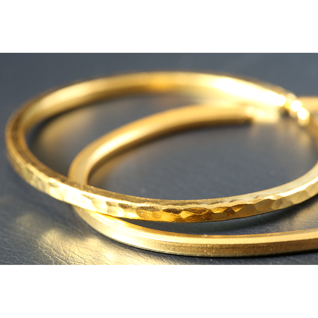 close up of two gold bangles placed on a dark surface one with a hammered texture and the other with a smooth matte finish